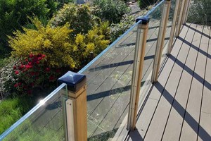 Raised decked balcony with glass panel railing overlooking a garden, featuring square plastic protective caps on the wooden balcony posts.