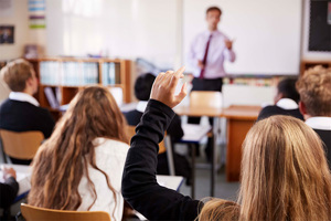 Student with hand raised in classroom, teacher at front of the class.